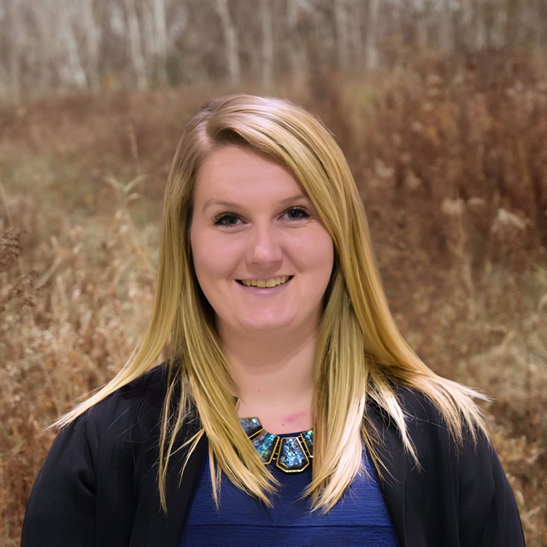 Smiling woman in a blue top and black cardigan standing outdoors with tall dry grass behind her