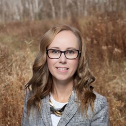 Portrait of a woman with glasses and wavy hair in a gray jacket, standing outdoors in a brown field