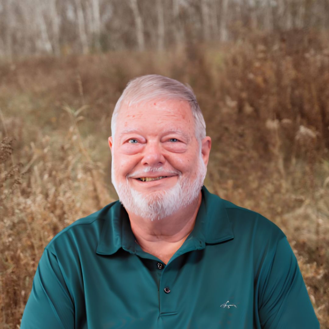 View Bio Smiling man in a teal polo shirt standing outdoors in a grassy field