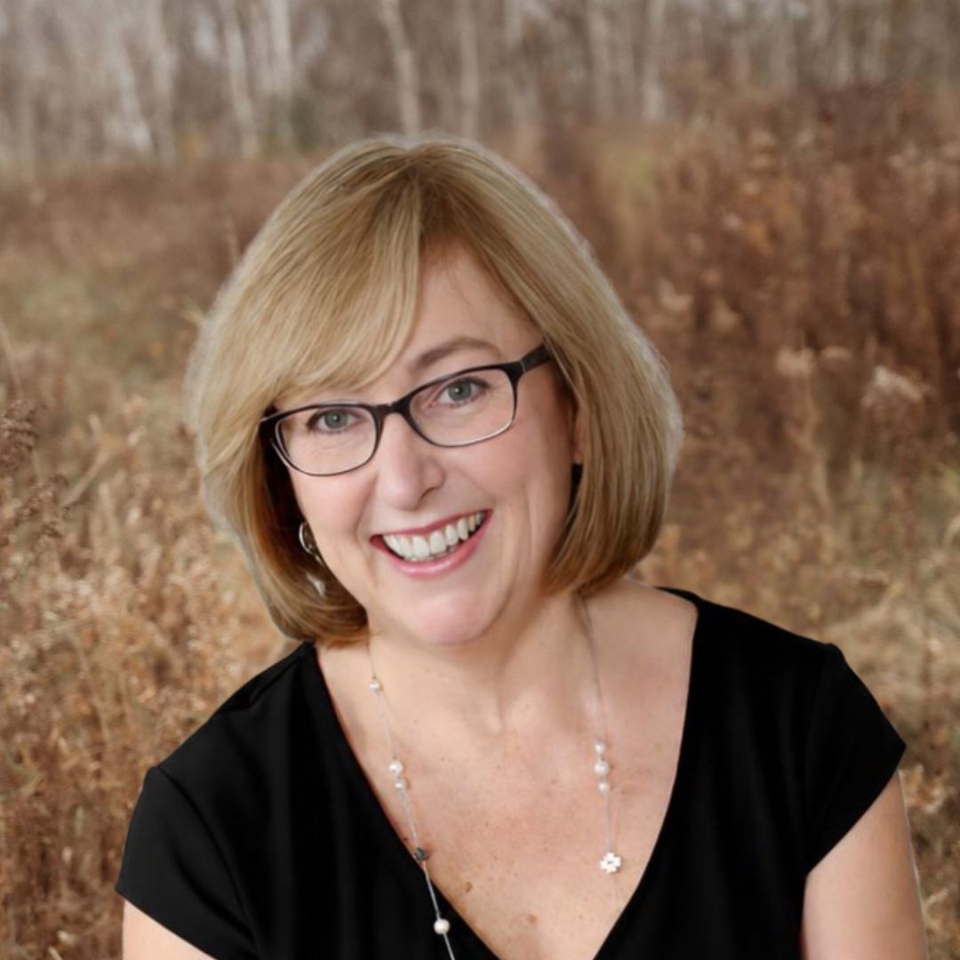 View Bio Smiling woman with glasses and blonde hair in a black top against a blurred outdoor background