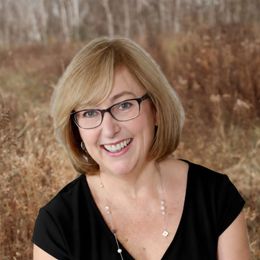 Smiling woman with glasses and blonde bob in a black top, posed against a blurred outdoor field background