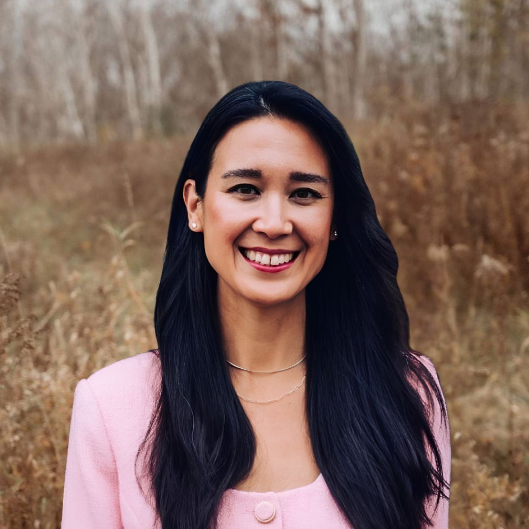 View Bio Smiling person in a pink top standing in a grassy field with tall, dry plants in the background