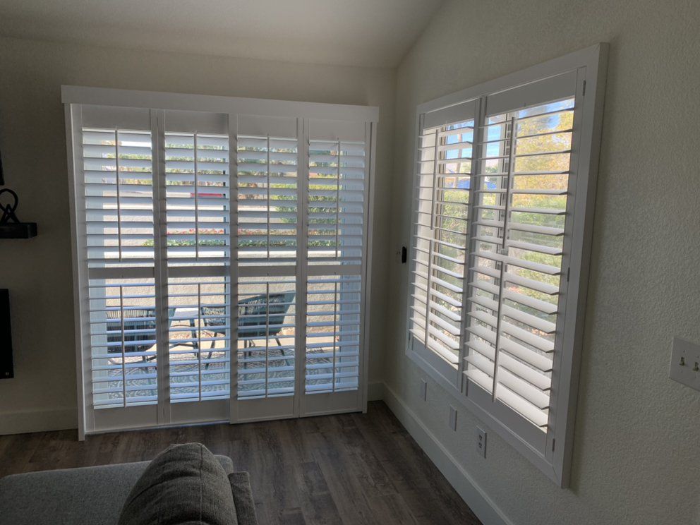 Interior view: White shuttered windows and a sliding glass door, allowing light and a view into a room with wood flooring.