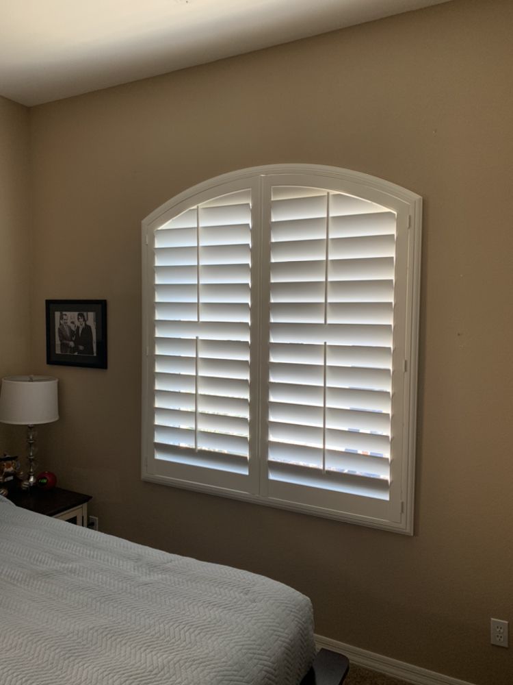 White shutters cover an arched window in a room with tan walls. A bed and nightstand are visible.