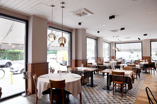Interno di un ristorante con tavoli apparecchiati per cenare, ampie finestre e lampade a sospensione.
