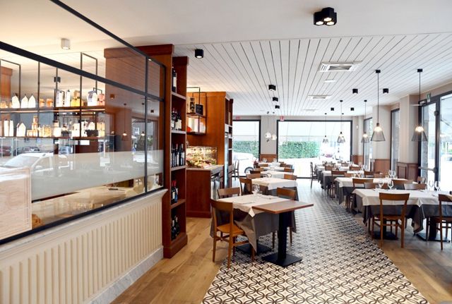 Interno di un ristorante con tavoli apparecchiati per cenare, pavimento a scacchiera e finestre alte.