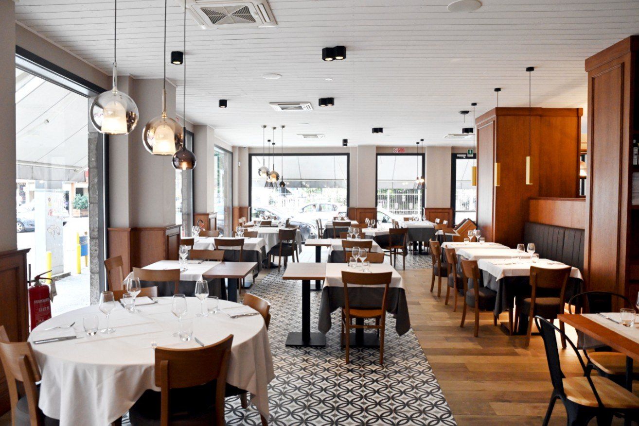 Interno di un ristorante con tavoli apparecchiati per cenare, pavimenti in legno, piastrelle a motivi e lampade a sospensione.