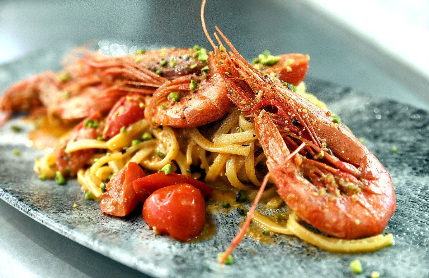 Piatto di pasta con gamberi grandi cotti, pomodori e guarnizione di pistacchi su un piatto grigio.