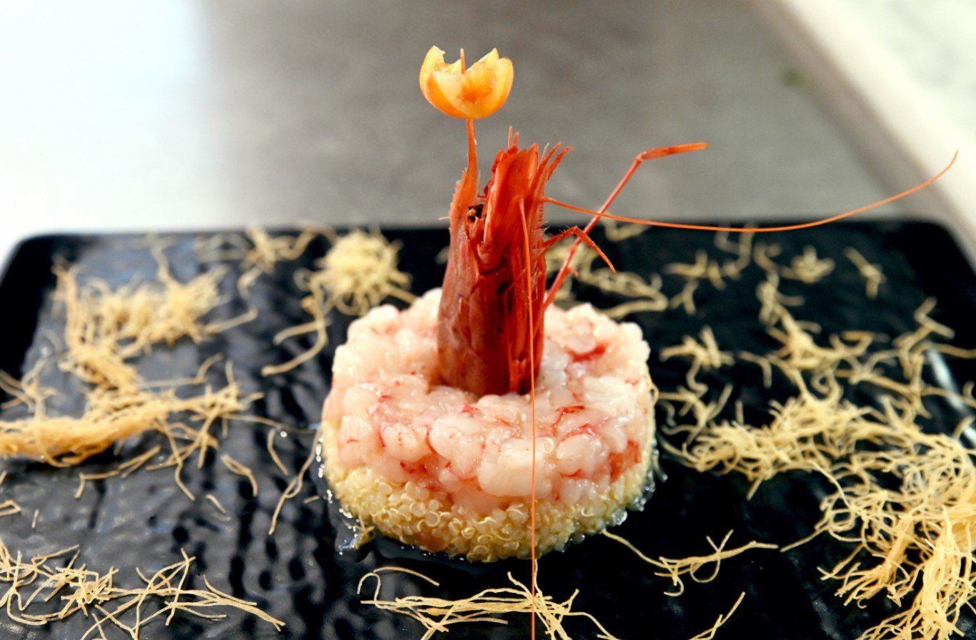 Piatto di gamberi, gamberi e quinoa in cerchio, guarnito con un gambero e un fiore, su un piatto nero, con noodles decorativi.