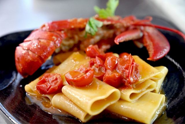 Piatto di pasta e aragosta con pomodori arrostiti, su un piatto nero.