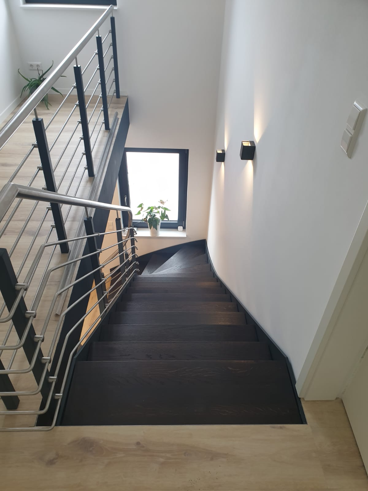 Modernes Treppenhaus mit dunklen Stufen, Metallgeländer, weißen Wänden und einem Fenster im Treppenabsatz.