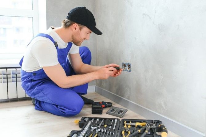Electrician in blue overalls installing an electrical outlet on a wall with tools spread on the floor.