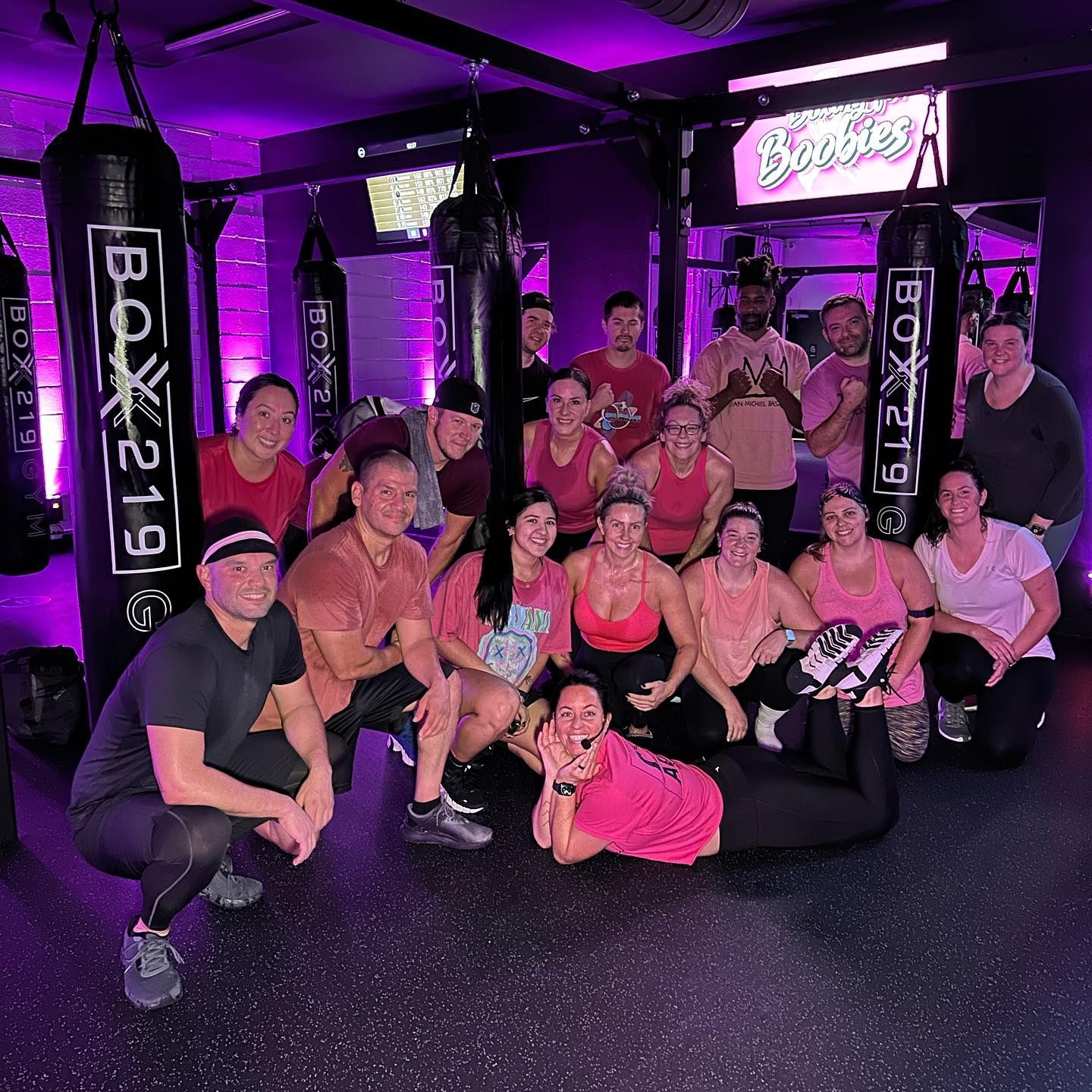 Group of people in a boxing gym wearing pink shirts pose in front of punching bags.