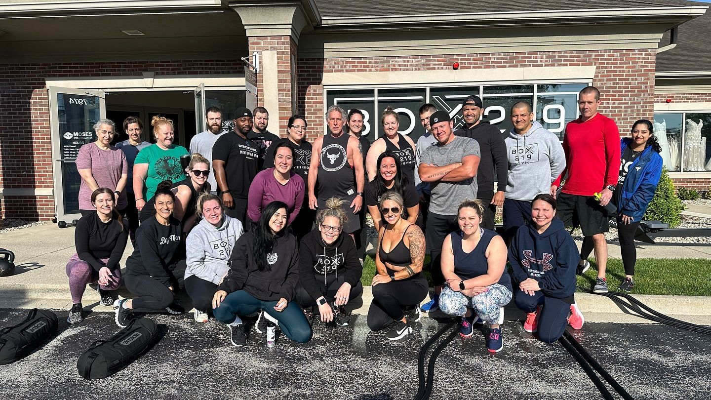 Group of people posing outside a fitness studio, some kneeling. Ropes and weights visible.