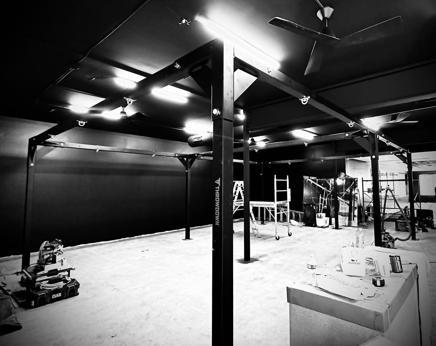 Black and white shot of an empty industrial-looking space, with metal beams, overhead lighting, and equipment along walls.