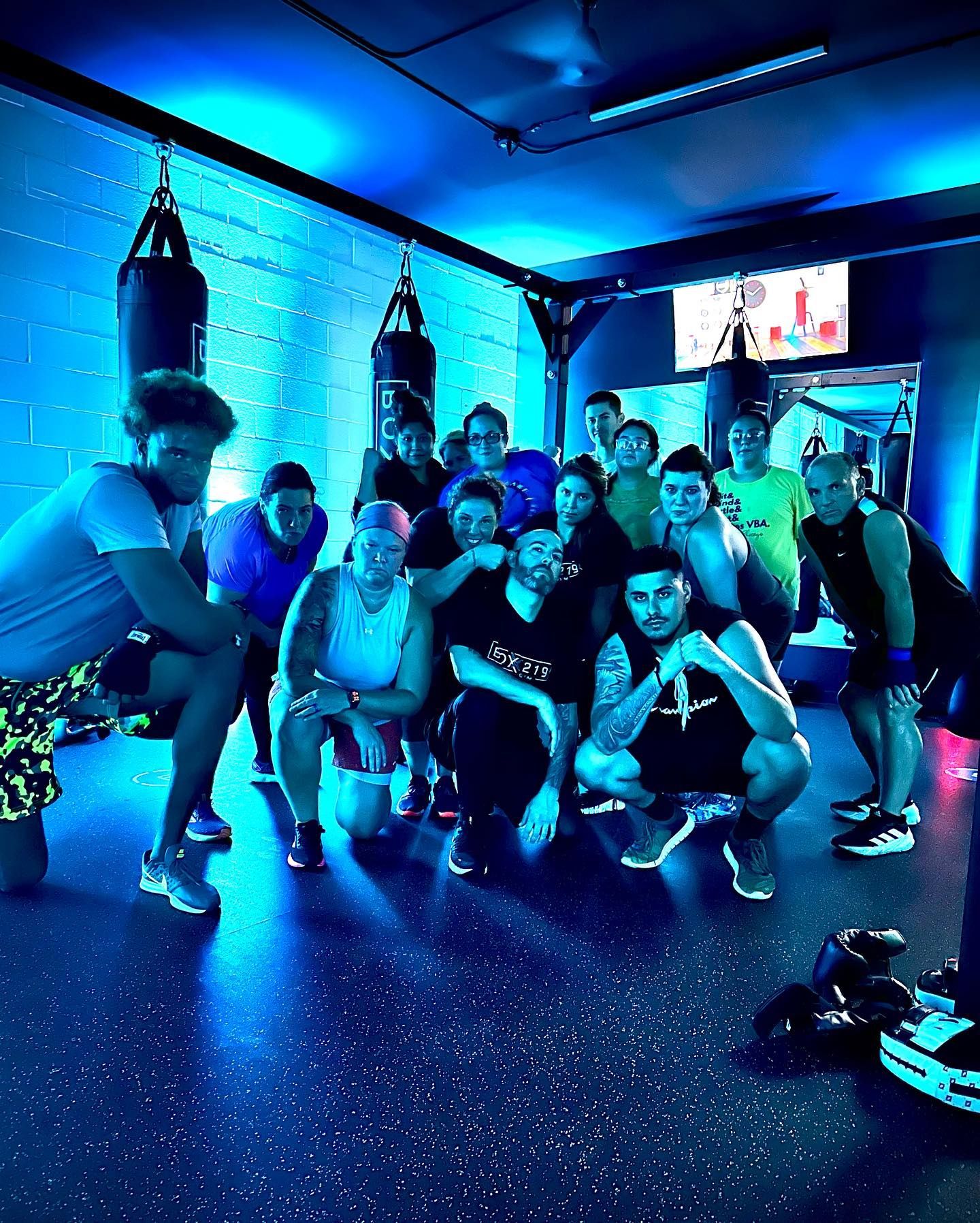 Group of people in a boxing gym, posing near punching bags. Blue lighting.