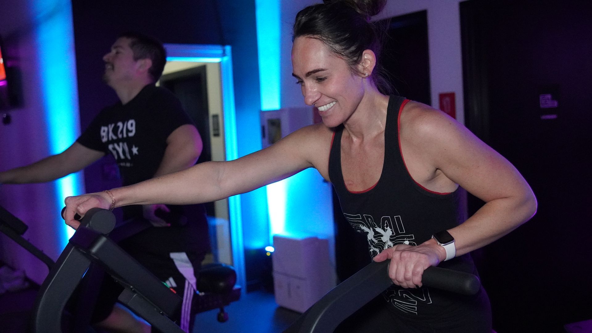 Woman and man working out on cardio machines, inside a gym with blue lighting.