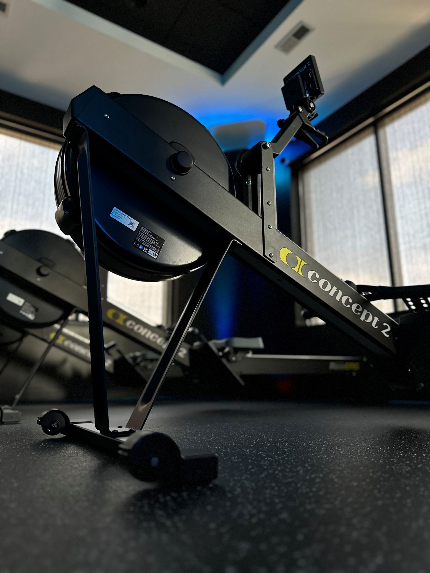 Black Concept2 rowing machine on a gym floor, with another in the blurry background, against a window.