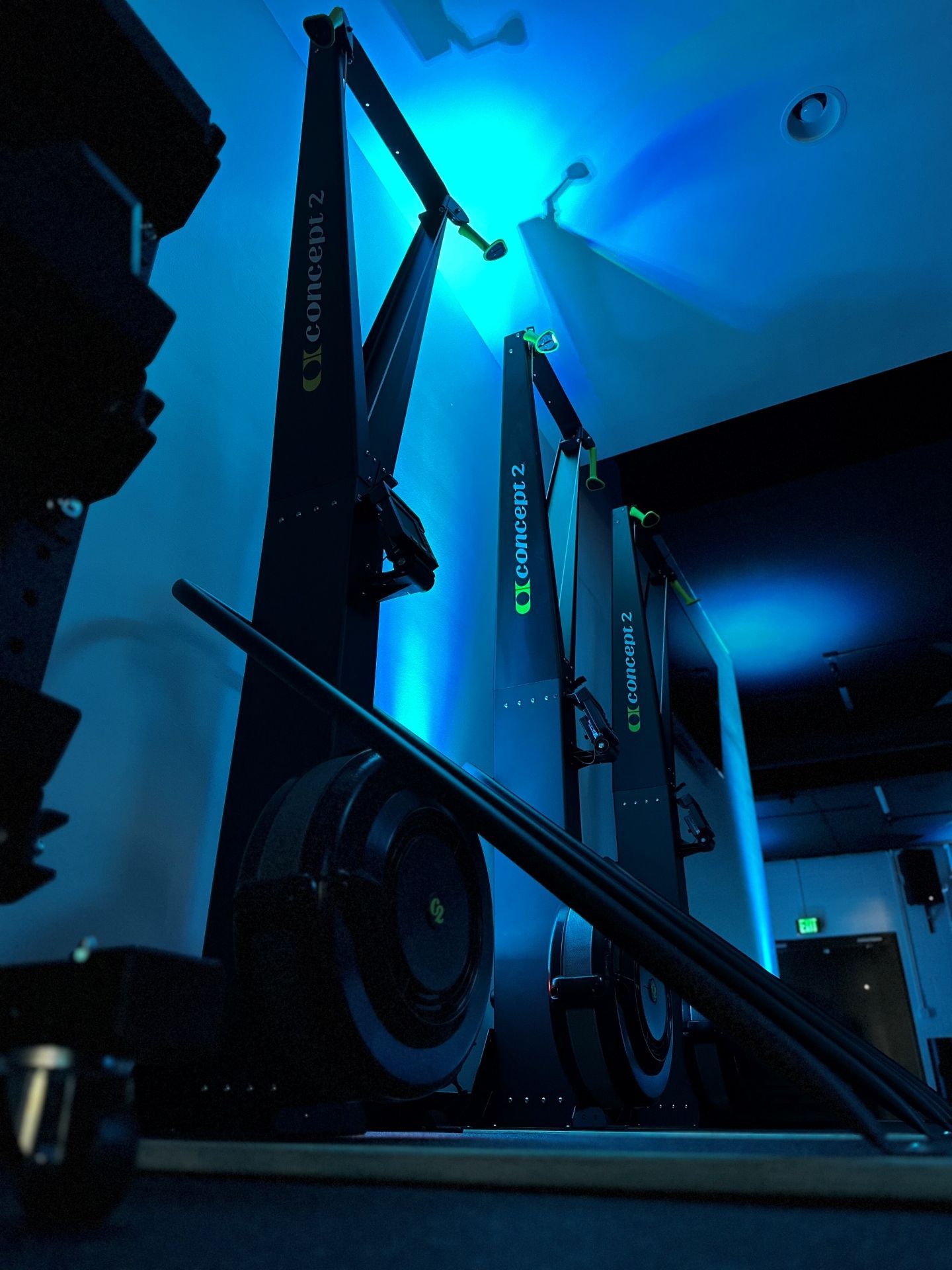 Two Concept2 rowers in a blue-lit gym. Oars rest in foreground.