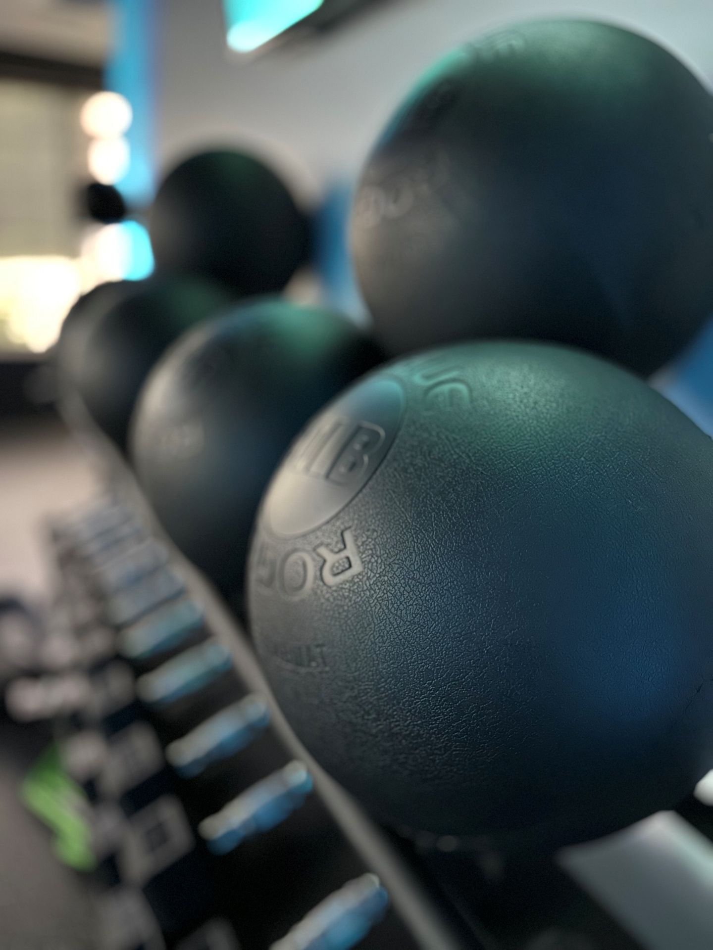 Black medicine balls stacked on a rack in a gym setting, with dumbbells in the blurred background.