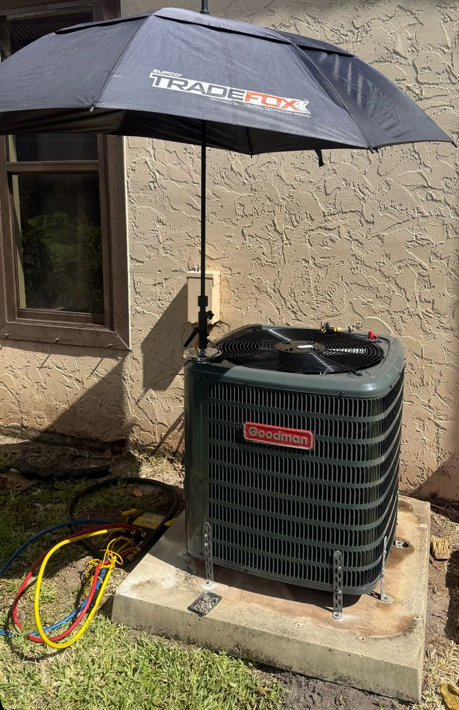 Outdoor HVAC unit with a black sun umbrella shade and colorful hoses beside a stucco wall.