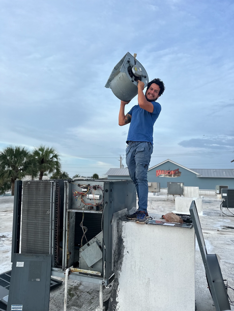 Man standing on rooftop lifting a large HVAC unit under a cloudy sky