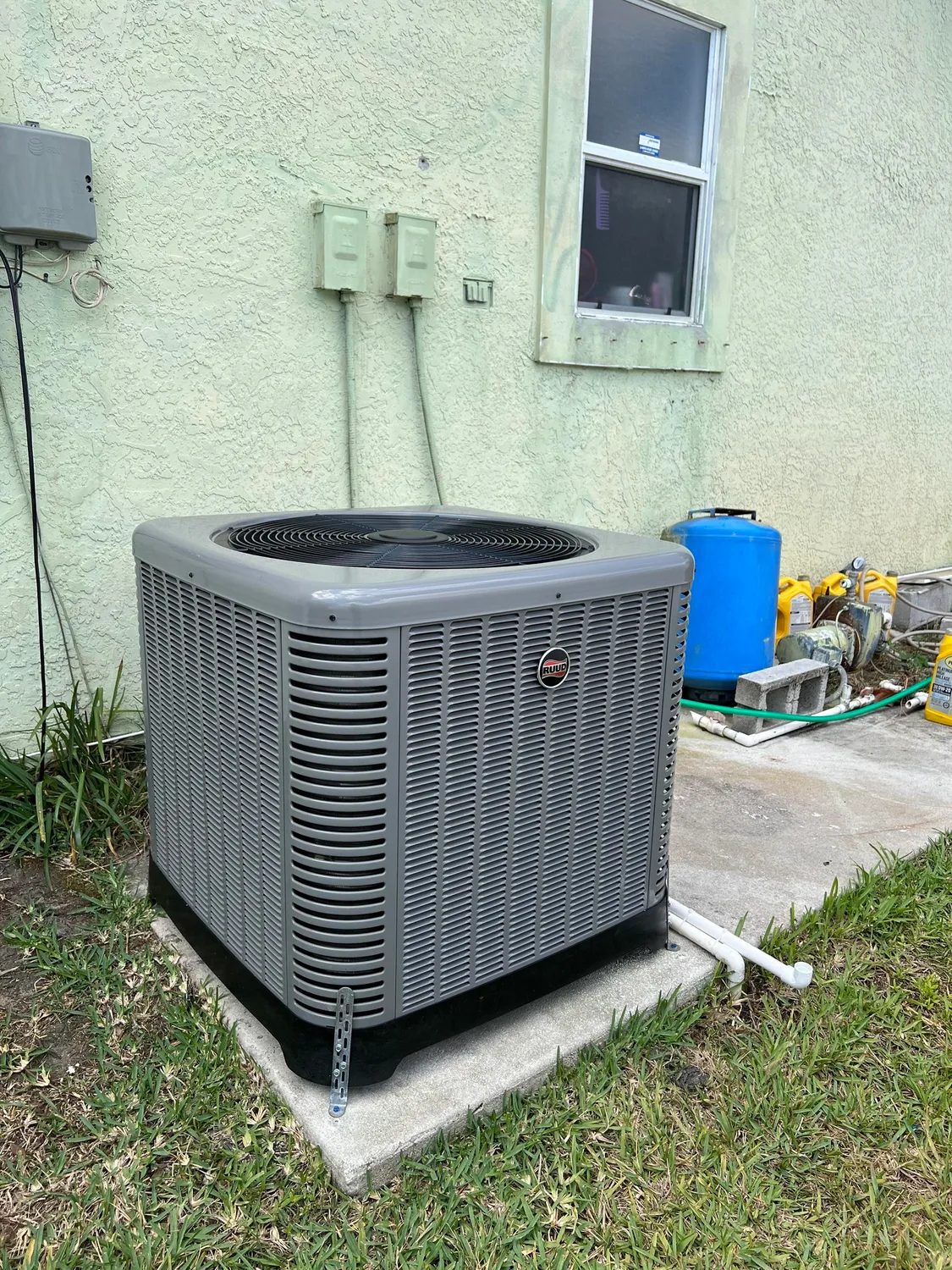 Outdoor HVAC unit beside a house wall, with a blue barrel and utility items nearby.