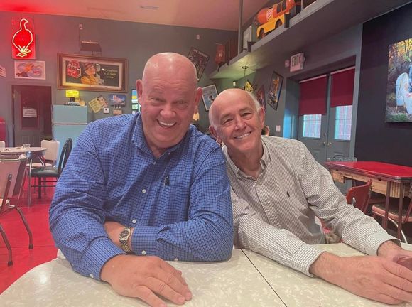 Two smiling people sit at a table in a red-lit restaurant, leaning close together.