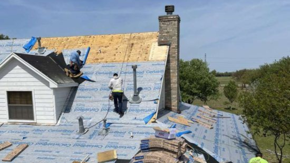 Roofers working on a house with a partially completed roof and a brick chimney under a blue sky.