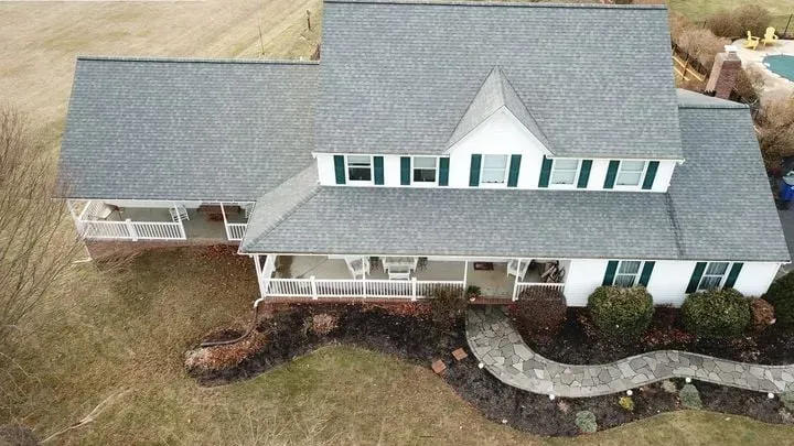 Aerial view of a white house with a gray roof and green shutters, surrounded by a lawn and walkway.