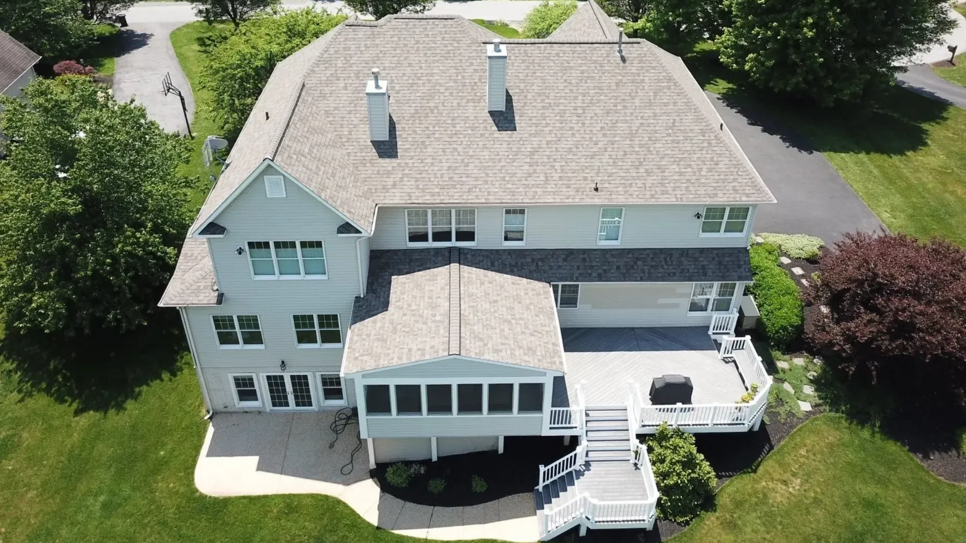 Aerial view of a two-story light gray house with a large deck and gray roof replacement. Green lawn surrounds the house.