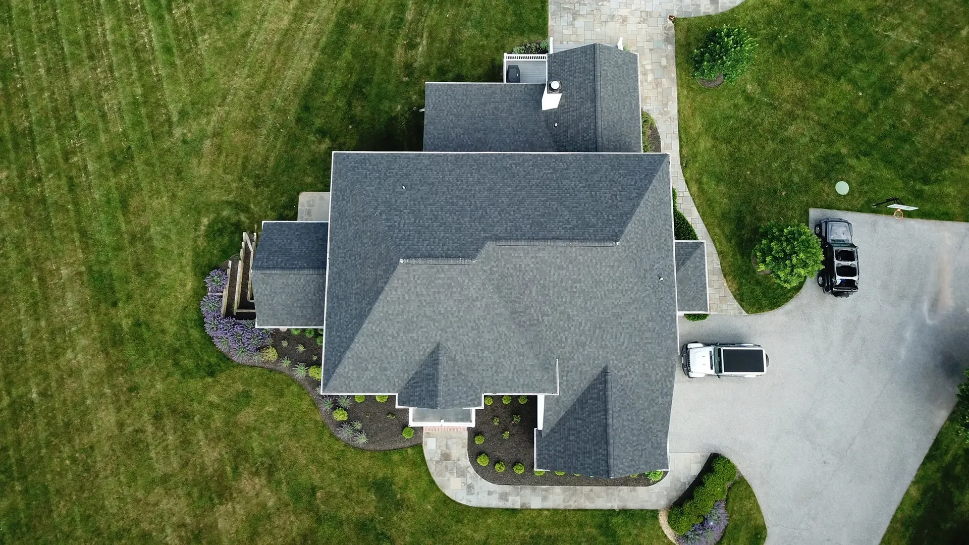 Overhead view of a house with a gray roof, surrounded by green grass and a paved driveway with cars.