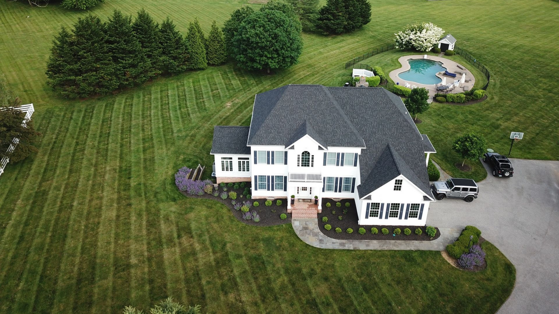 White house with dark roof and large lawn, driveway, and pool. Roof replacement project
