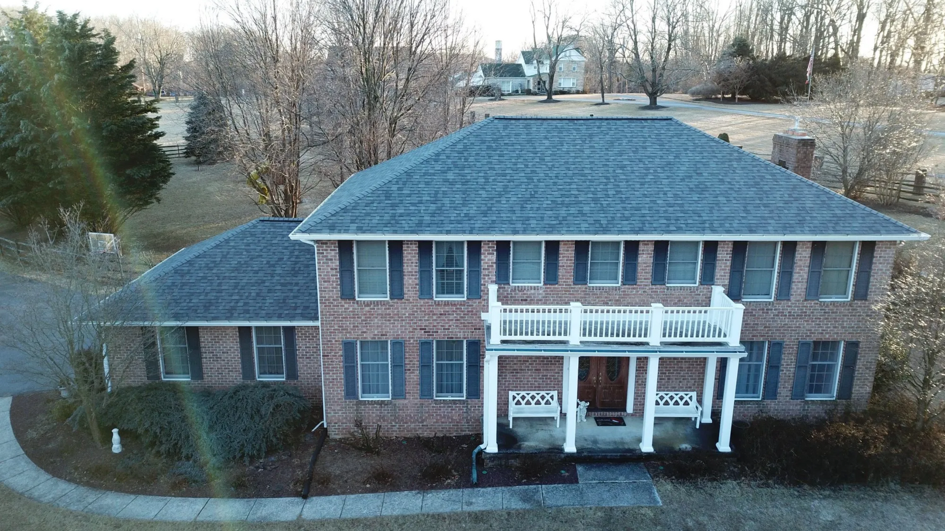 Two-story brick house with blue shutters and a white balcony, set in a grassy yard with bare trees.