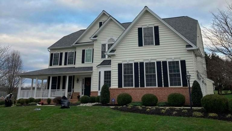 Two-story cream house with black shutters, red brick base, porch. White siding installation.