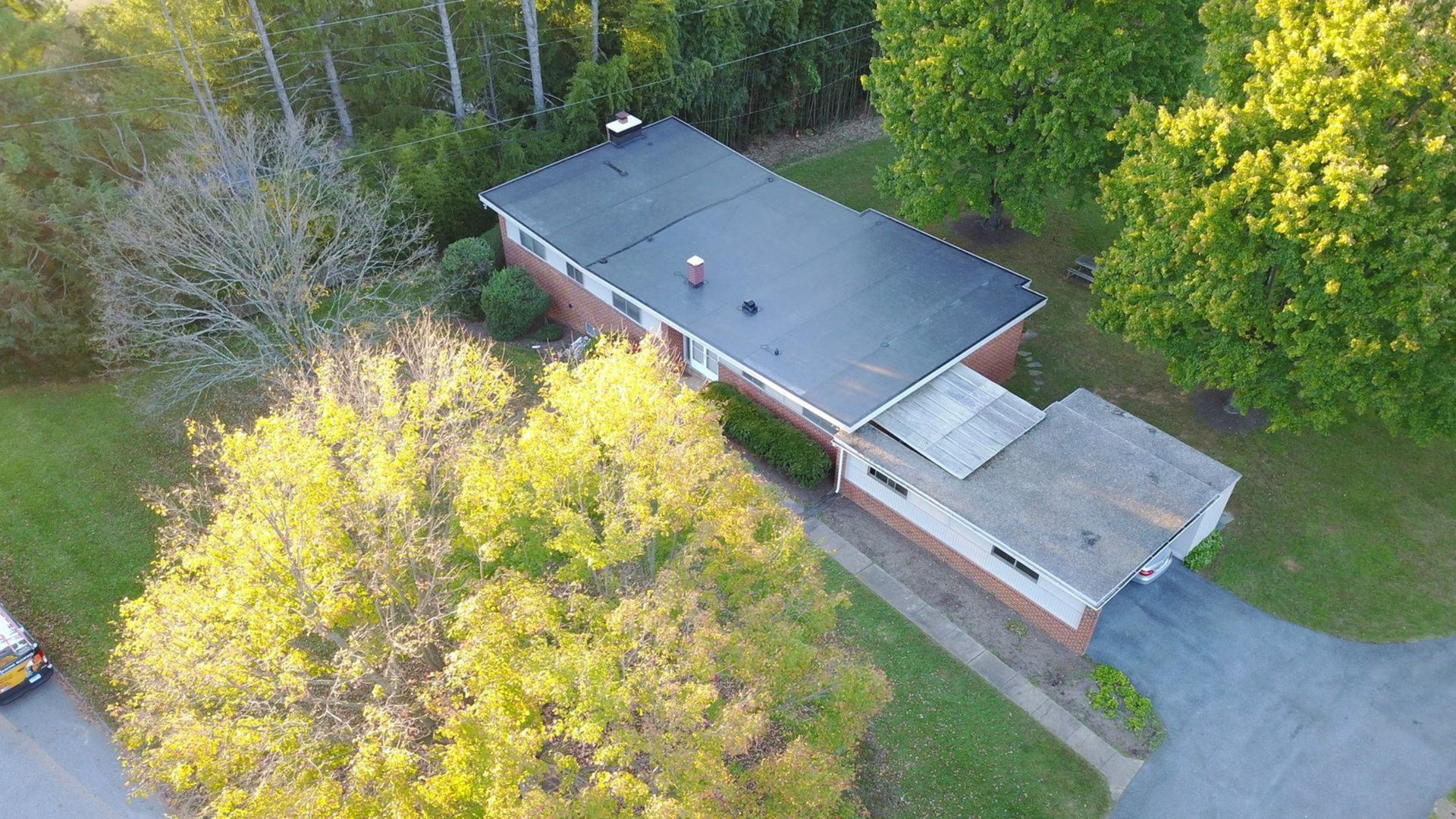 Aerial view of a single-story house with a flat black roof surrounded by trees and a driveway.