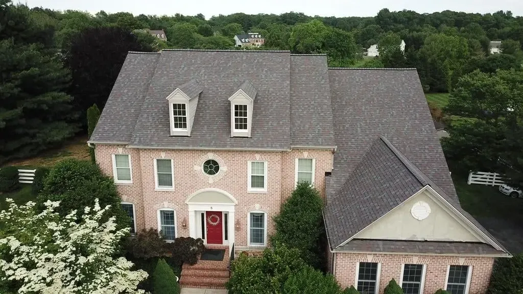 Two-story brick house with a dark gray roof, three dormers, and a red front door, surrounded by greenery.