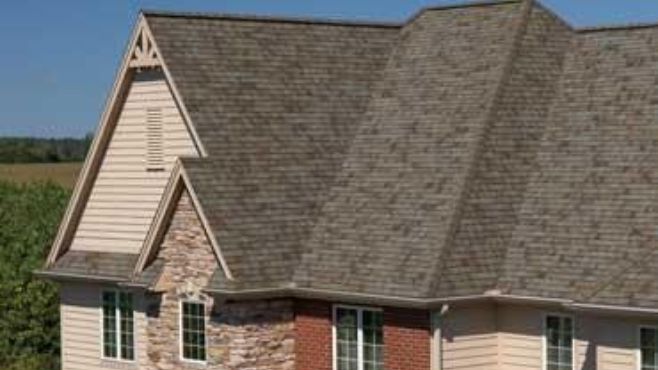 Tan house with brown shingled roof, windows, brick and stone accents, blue sky.