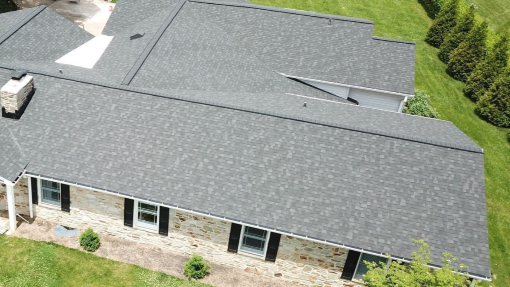 Grey shingled roof of a house with stone and black shutters, set in a grassy yard.