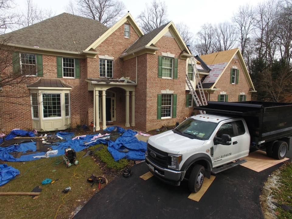 A brick house with roof damage, blue tarp on the lawn, and a dump truck in the driveway.