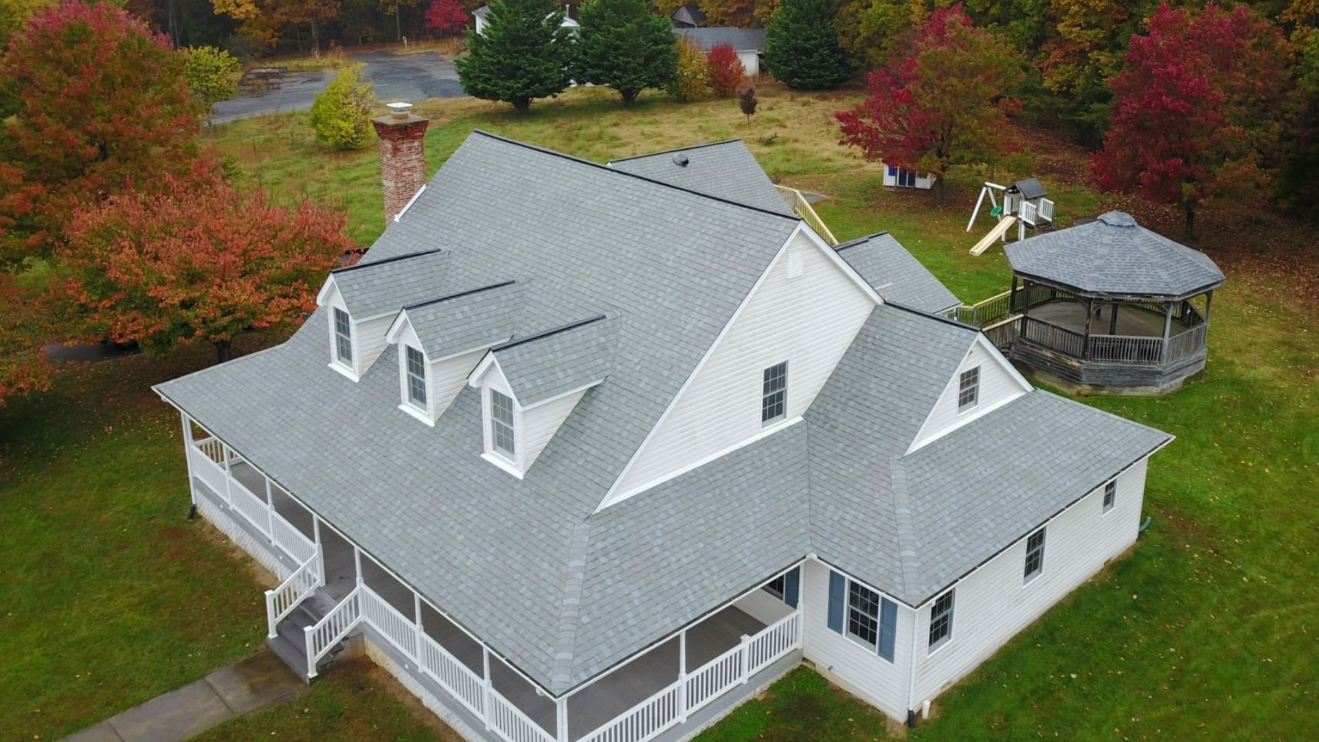 Gray-roofed house with white siding and porchand dormers, surrounded by green grass.. Fourseasonsroofing project