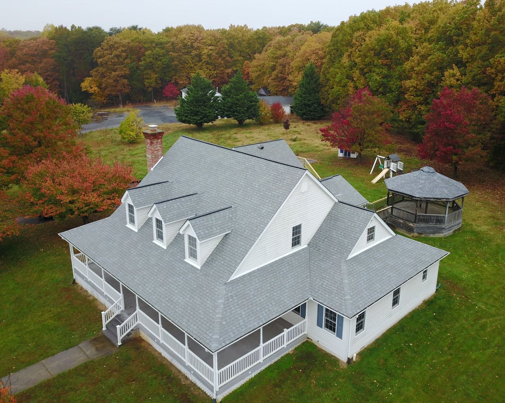 Aerial view of a gray-roofed house, surrounded by trees with fall foliage and a gazebo. Roof replacement by Four Seasons