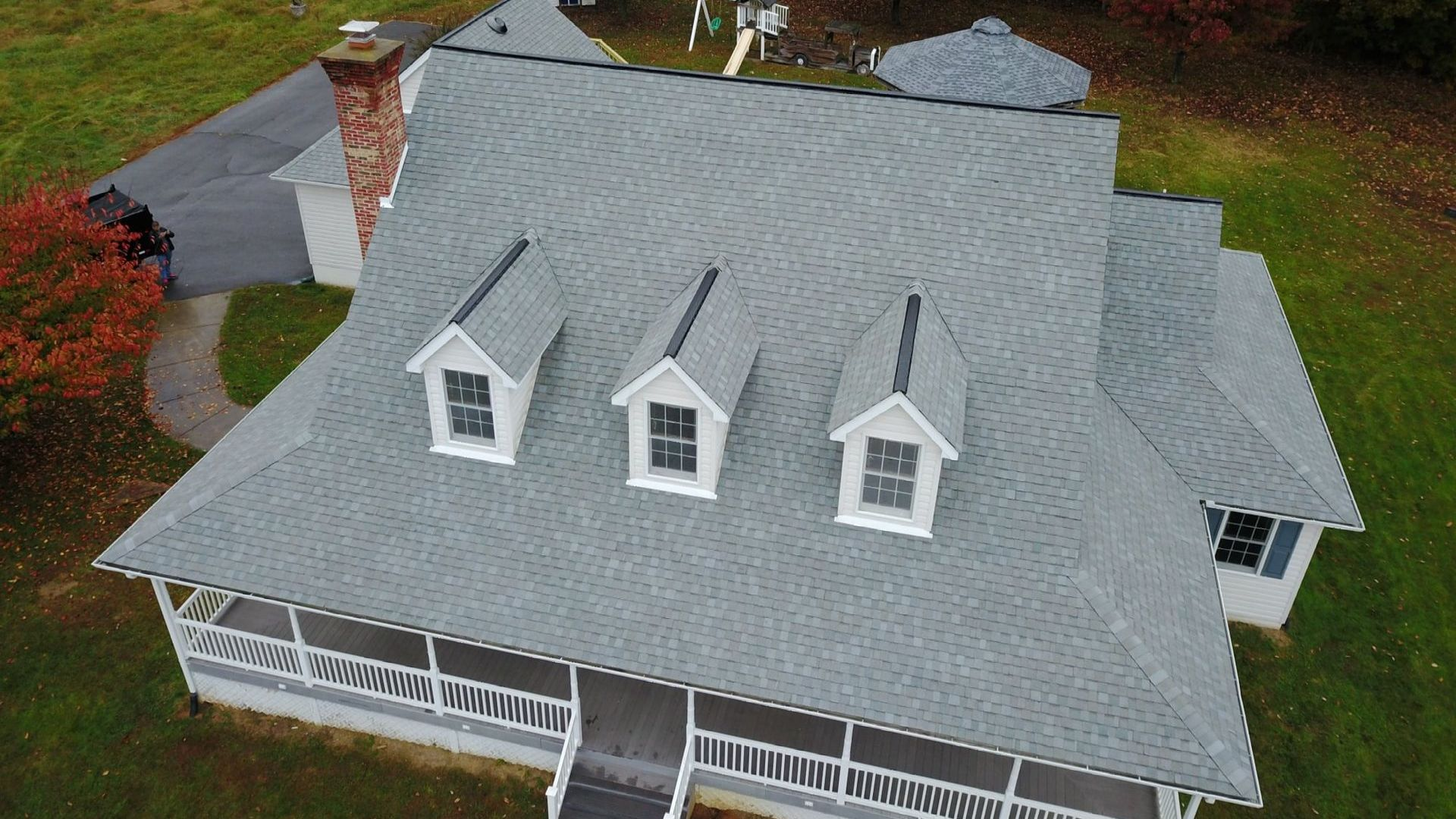 Gray-roofed house with three dormers, porch, and a winding driveway, set in a grassy area with some red-leaved trees.
