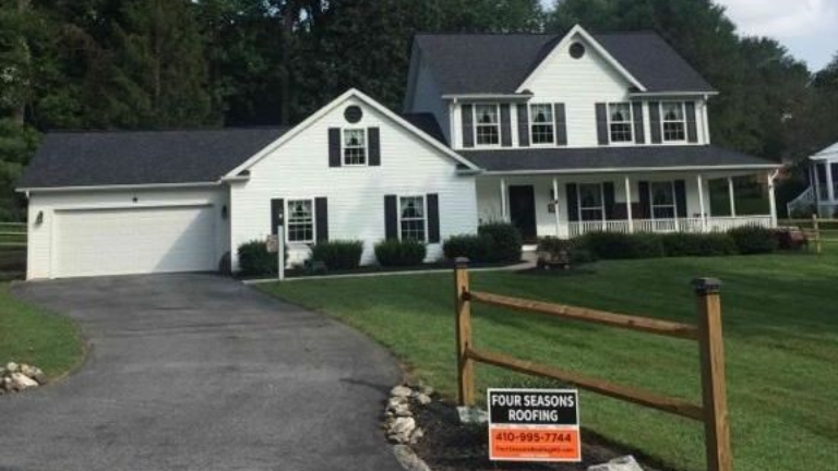 White two-story house with dark gray roof, black shutters, and driveway; sign in front.