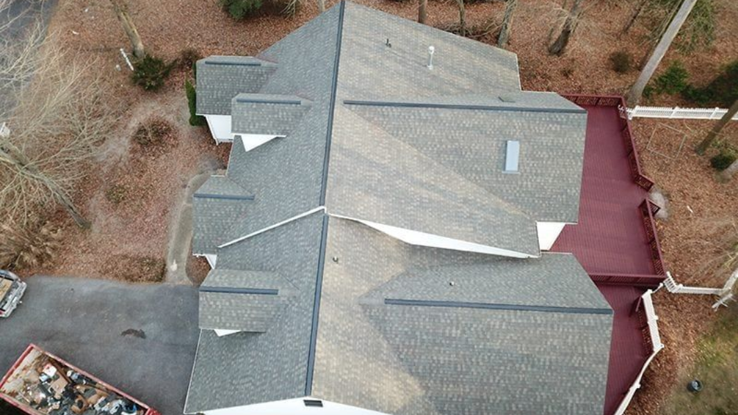 Aerial view of a gray-shingled house with multiple roof sections and a red-paved driveway.