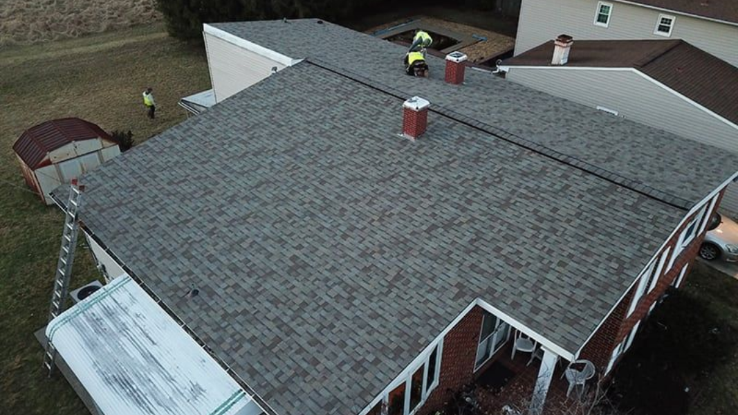 Workers on a roof with two chimneys; a ladder and a small shed in the yard.