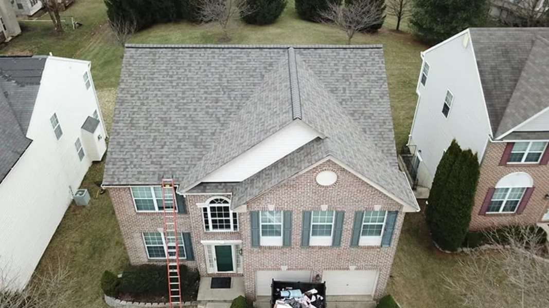 Overhead view of a two-story brick house with gray roof, green shutters, & a ladder leaning  to get to the roof