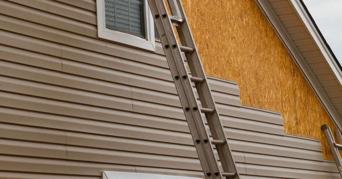A ladder leans against a house with beige siding near an upper window and yellow stucco wall.