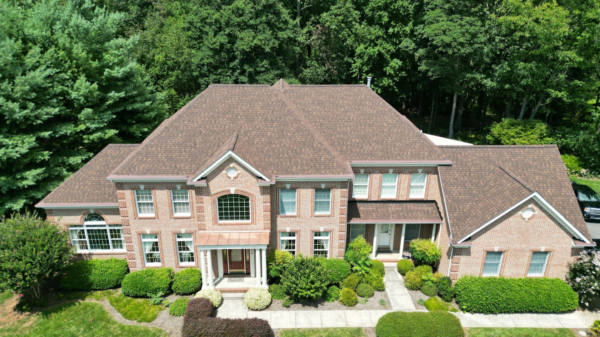 Large brick house with brown roof and manicured landscaping. Green trees in background. Roofing replacement service.