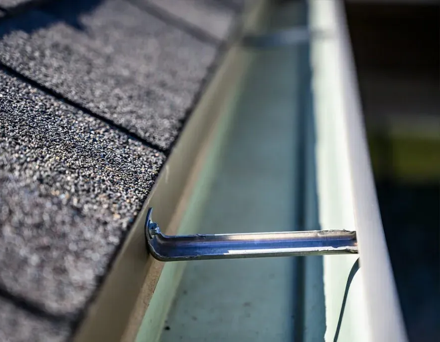 Close-up of a roof gutter, silver metal bracket, attached to the roof shingles, and the edge of the roof.
